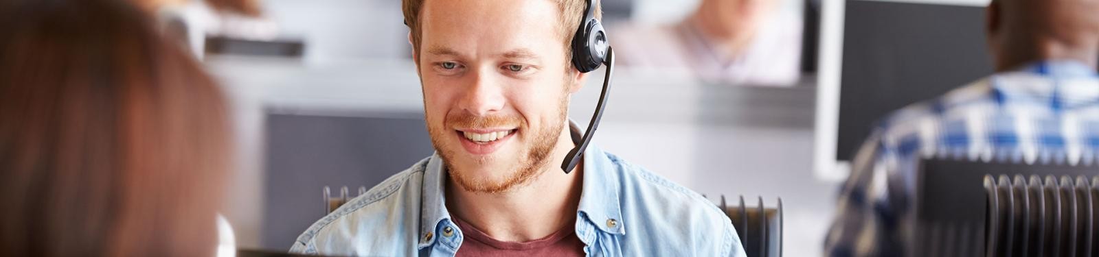 Sprechzeiten Ein Mann mit Headset sitzt in einem Großraumbüro am Schreibtisch.