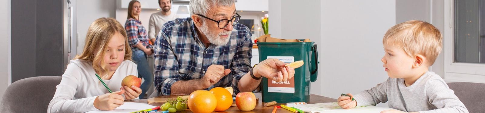 Ein älterer Mann sitzt mit zwei Kindern am Esstisch; auf dem Tisch befindet sich Obst und ein Bioabfallsammelbehälter