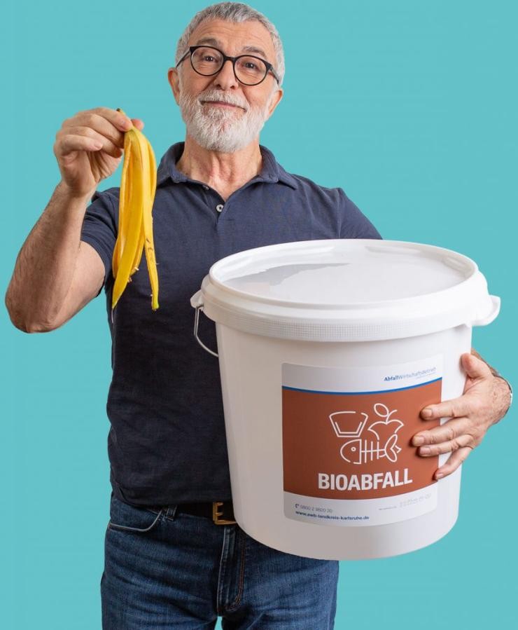 Ein Mann hält mit der rechten Hand eine Bananenschale hoch mit dem linken Arm umfasst er eine weißen Eimer für Bioabfall. 