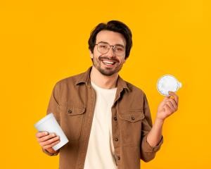Ein Mann steht vor einer gelben Wand und hält glücklich einen Joghurtbecher in der einen und den Deckel davon in der anderen Hand