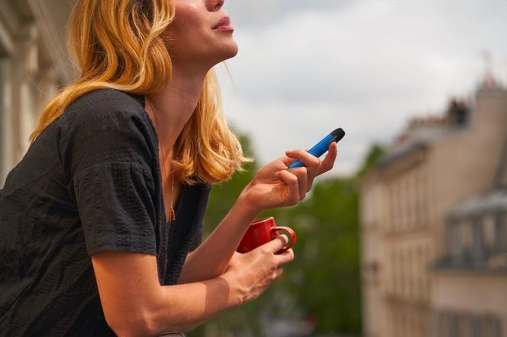 Eine Frau im T-Shirt steht  auf einem Balkon, raucht eine blaue Einmal-E-Zigarette und hält in der anderen Hand eine rote Tasse.