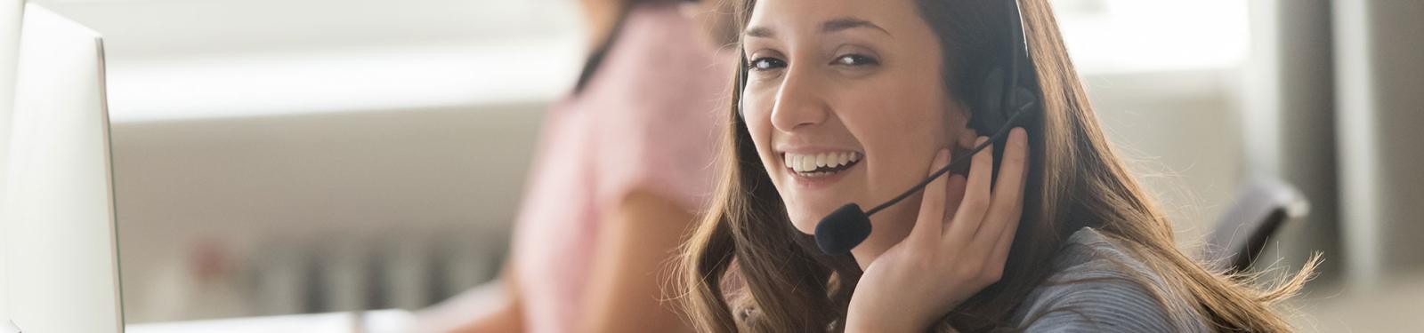 Unsere Services Eine junge Frau mit Headset sitzt am Schreibtisch und lächelt in die Kamera.