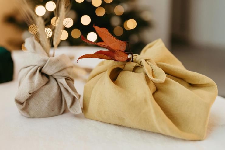 Zwei Geschenke sind in einem Stofftuch, in der Furoshiki-Technik, eingewickelt. Im Hintergrund schimmert ein geschmückter Weihnachtsbaum. Nachhaltige Geschenkverpackung aus einem Stofftuch in der Furoshiki-Technik.