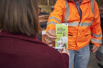 Ein Mann mit Arbeitskleidung überreicht einer Frau einen Flyer zu Bioabfall