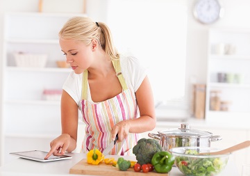 Eine Frau liest in der Küche in einem IPad. Vor ihr steht ein Schneidebrett und viele verschiede Gemüsesorten