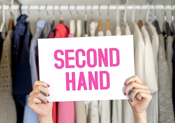 Eine Person hält ein Schild mit der Aufschrift "Second Hand" hoch, im Hintergrund befindet sich ein Kleiderständer