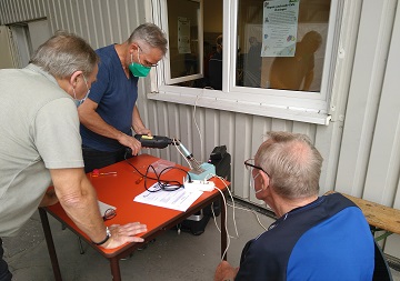 Ein Mann repariert ein Elektrogerät an einem Tisch, zwei Männer schauen zu